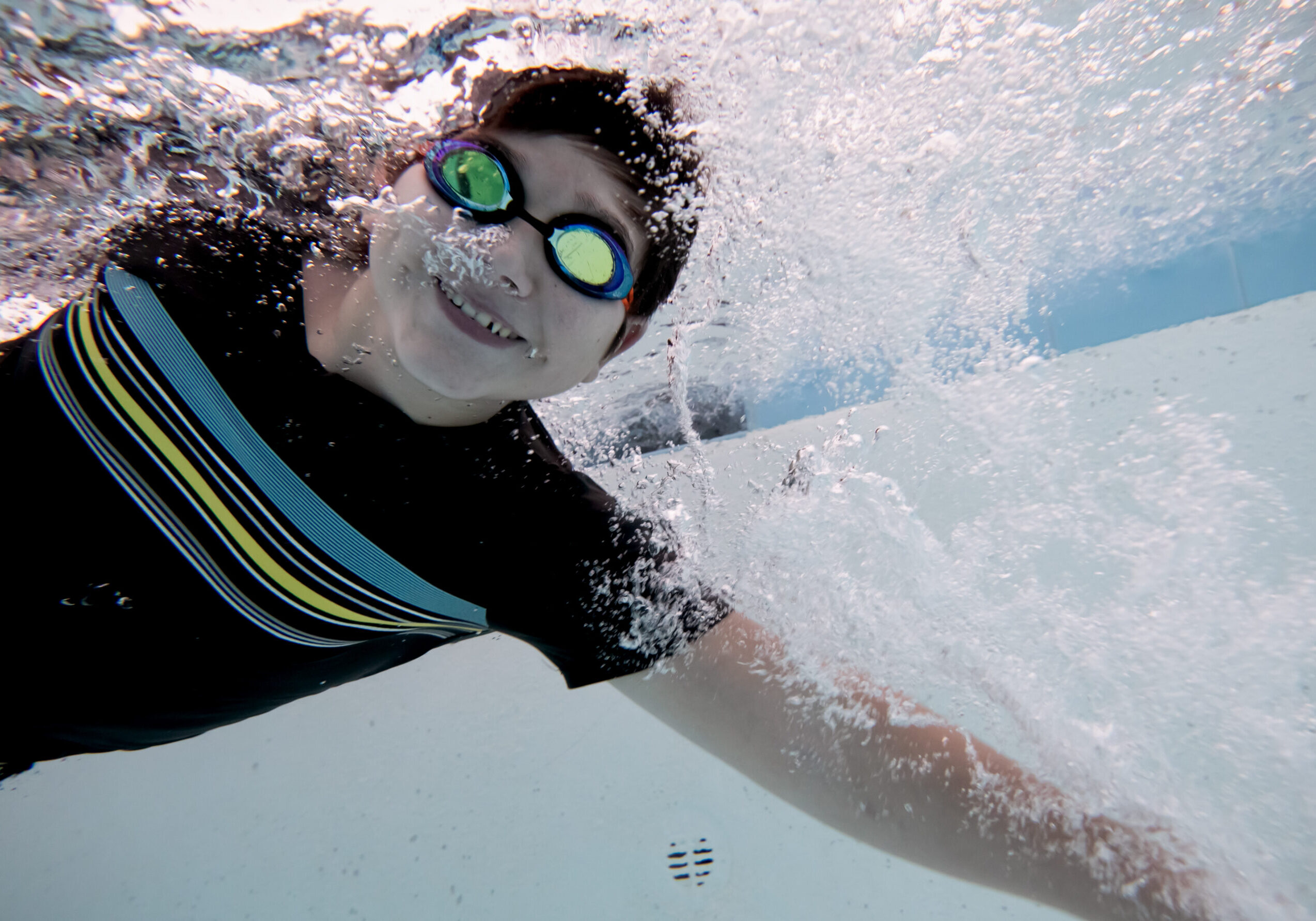 young boy swimming underwater, looking at the camera and smiling as he feels empowered and comfortable in the pool.